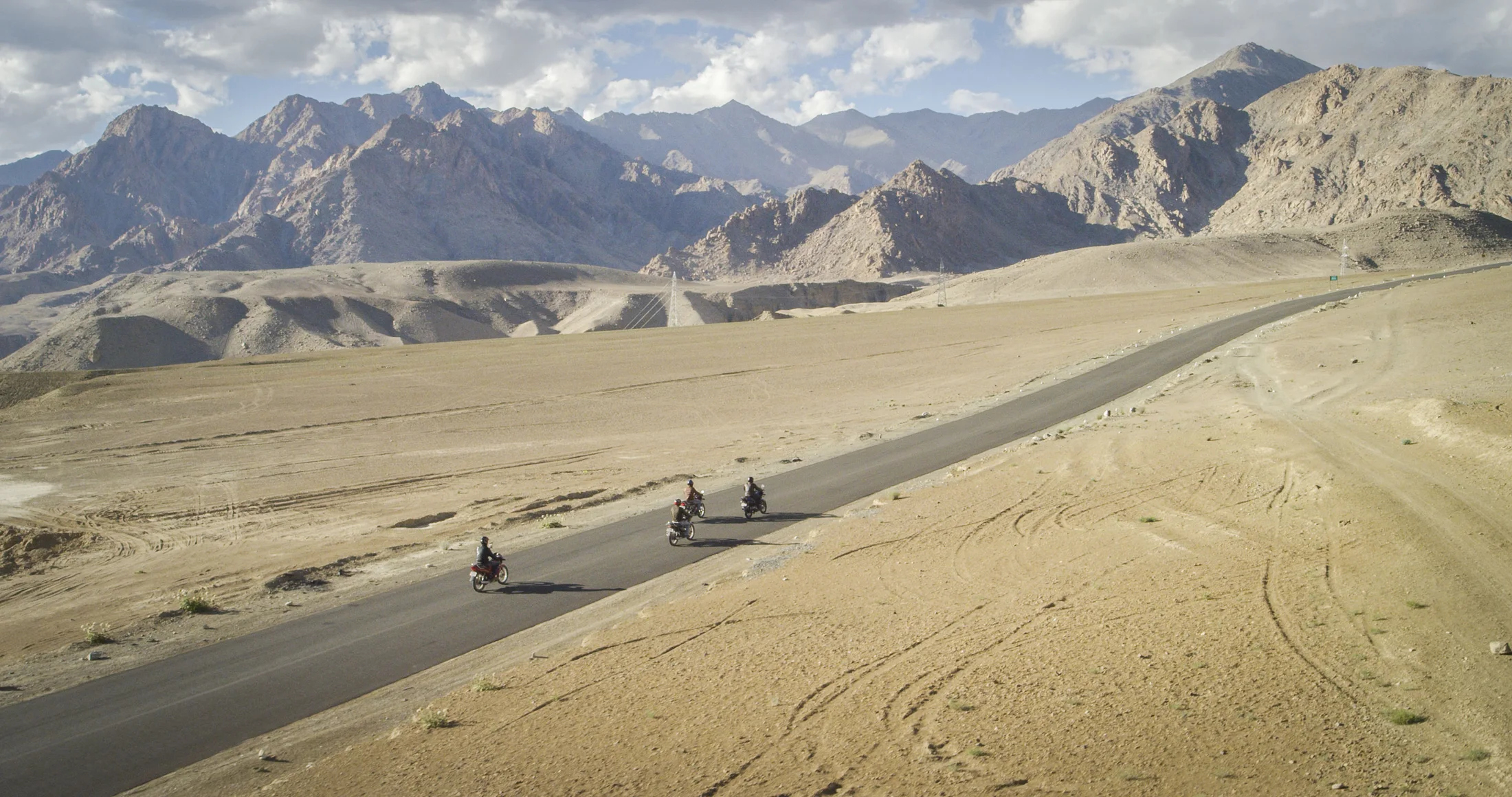 Four motorcyclists riding together along a wide road through Ladakh's high-desert landscape